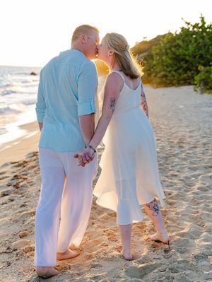 Couple kissing while holding hands on a tropical beach at golden sunset, dressed in white and light blue.