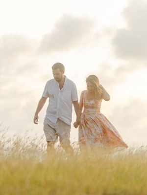 Couple holding hands walking through golden field at sunset, woman in floral dress, man in casual white shirt.