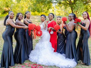 Bride in white mermaid gown poses with bridesmaids in black dresses holding red bouquets outdoors.