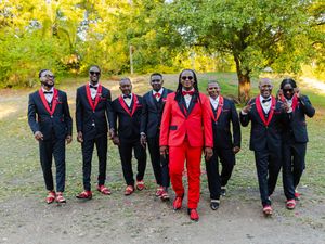 A groomsmen group in matching black tuxedos with red accents poses outdoors, with the groom in a bold red suit.