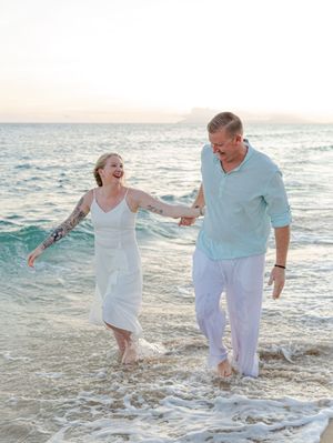 Joyful couple laughing and walking through ocean waves on a beach at sunset, holding hands barefoot.
