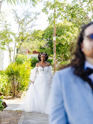 Bride in off-shoulder lace ball gown with floral crown walks toward groom in light blue suit at outdoor wedding.