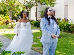 Bride in white off-shoulder gown and groom in light blue suit stand outdoors during a sunny wedding ceremony.