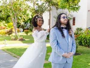 Bride in white off-shoulder gown taps groom in light blue suit during first look at tropical outdoor wedding venue.