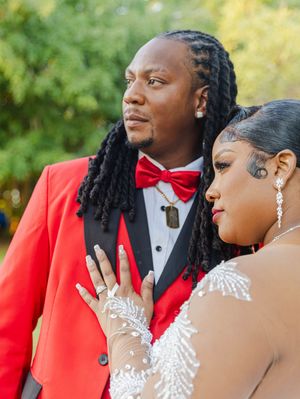 Black wedding couple posing outdoors, groom in red blazer with dreadlocks, bride in white beaded dress with henna hands.