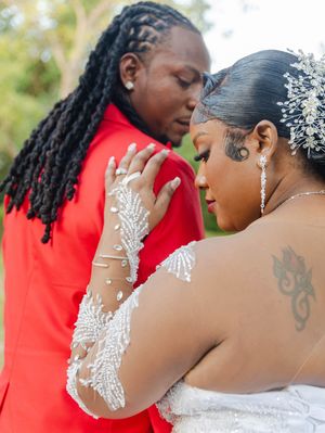 A bride in beaded lace gown and crystal headpiece shares an intimate moment with her groom in a red suit.