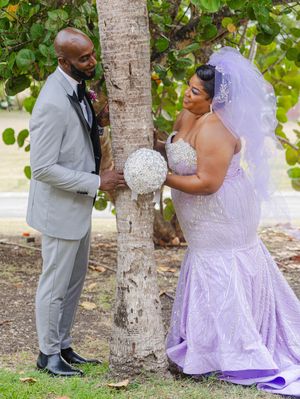 Bride in lavender mermaid gown with veil holds bouquet beside groom in gray suit near palm tree outdoors.