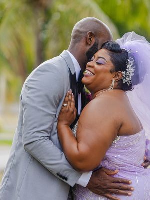 A joyful bride in a lavender gown and veil embraces her groom in a gray suit during their outdoor wedding celebration.