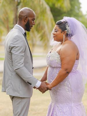A bride in lavender gown and veil holds hands with groom in gray suit during outdoor wedding ceremony.
