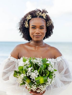 Bride with natural curly hair adorned with baby's breath flowers, holding a green and white bouquet by the ocean.