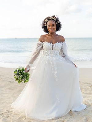 Bride in off-shoulder lace wedding gown with floral crown holds bouquet on sandy beach with ocean backdrop.
