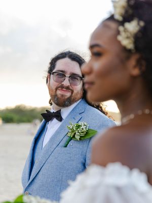 Groom in light blue suit with boutonniere smiles at bride in white dress with floral hair accessory at beach wedding.
