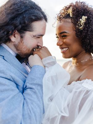 Interracial couple shares romantic moment as groom kisses bride's hand at outdoor beach wedding ceremony.