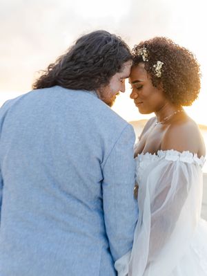 A couple in wedding attire shares an intimate forehead touch at sunset on a beach, bride in white off-shoulder gown.