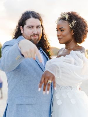 Newlywed couple showing off wedding rings, bride in white off-shoulder gown with floral hair, groom in blue suit at sunset.