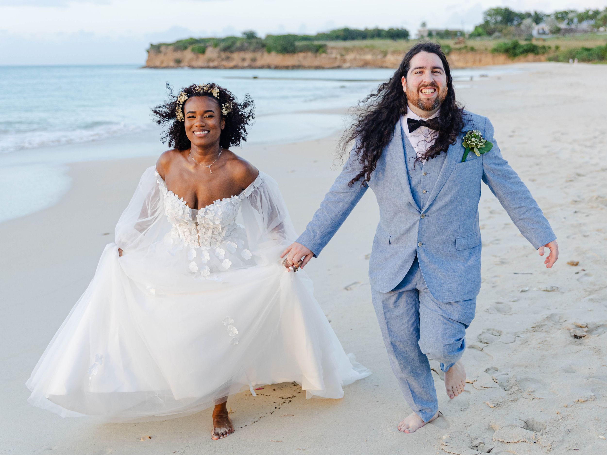 Joyful newlyweds in wedding attire running hand-in-hand on a sandy beach, bride in white gown, groom in blue suit.