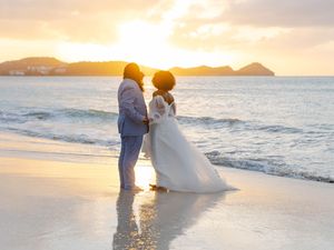 Bride and groom share a romantic moment on a sandy beach at golden sunset with ocean waves and mountains in the background.
