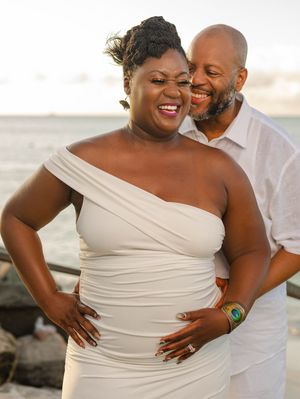 A joyful pregnant couple poses on a beach at sunset, woman in white one-shoulder dress, man in white shirt behind her.