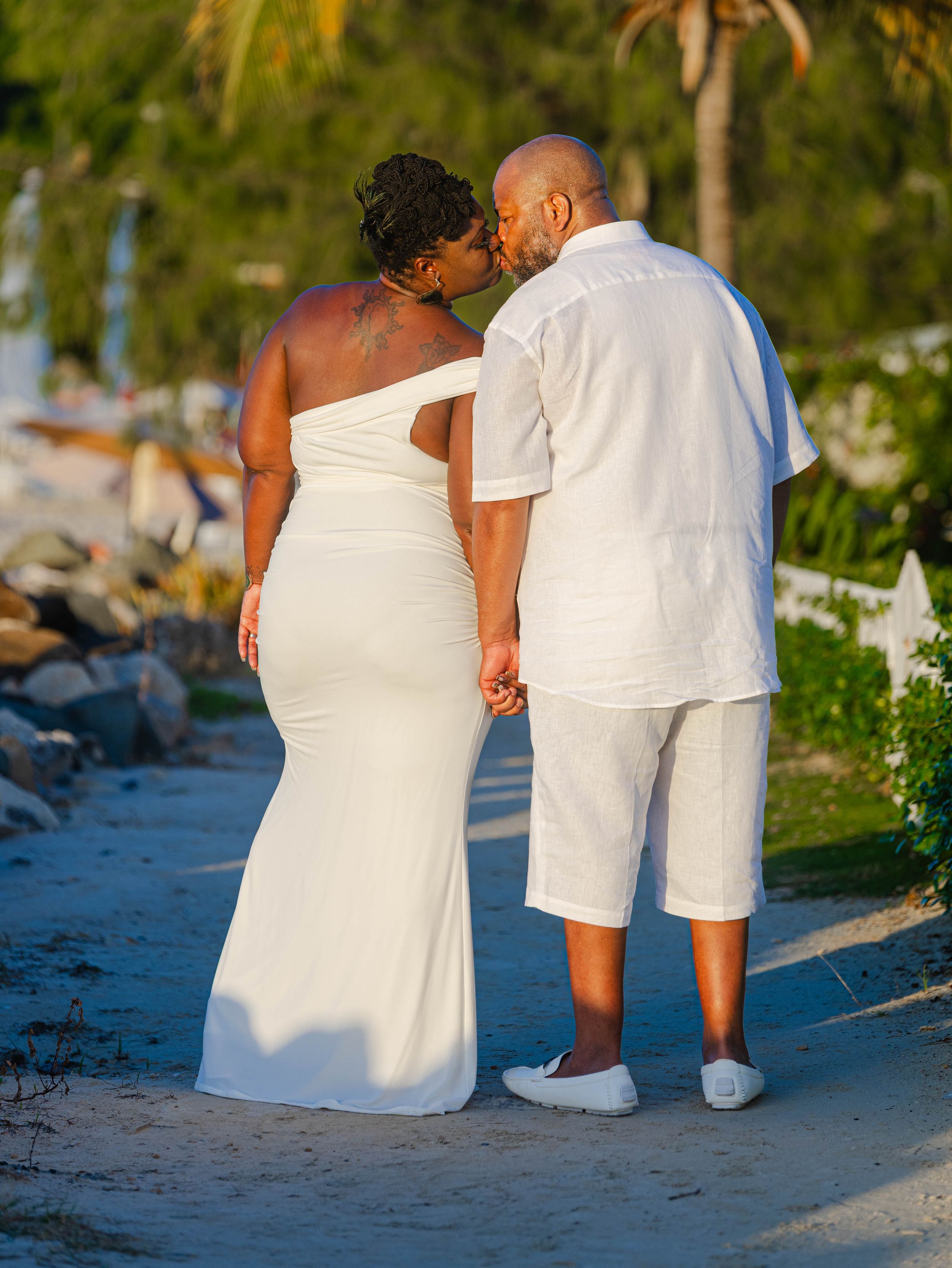 Couple in white outfits sharing a kiss on a sandy beach path at sunset, holding hands near tropical greenery.