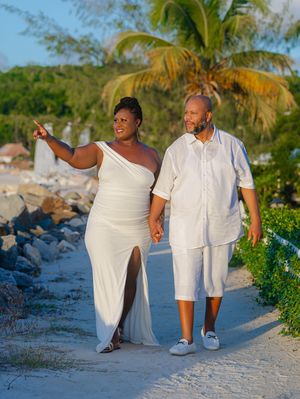 Couple in white outfits walking hand-in-hand on a tropical beach path lined with palm trees and rocks.