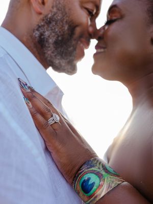 Couple sharing a romantic moment, woman wearing a diamond ring and peacock feather bracelet, backlit by golden sunlight.