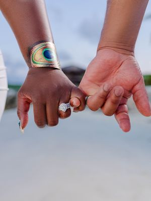 Close-up of couple holding hands showing diamond engagement ring and peacock feather bracelet on beach.