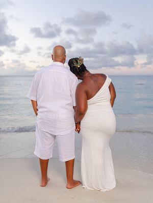 Romantic couple in white outfits holds hands while gazing at ocean sunset on tropical beach.