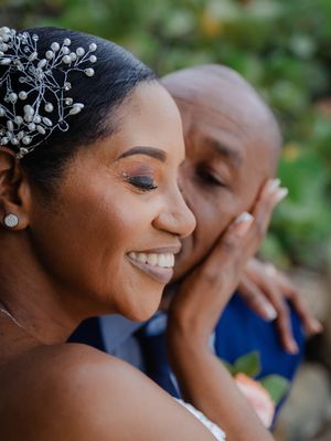 A joyful bride with floral hair accessory smiles as her groom tenderly kisses her cheek on their wedding day.