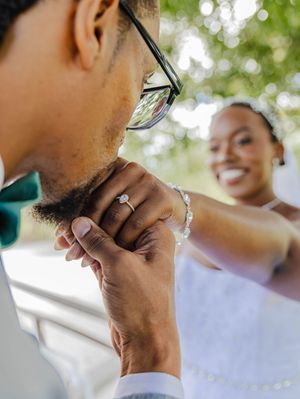 A bride smiles as she adjusts her groom's green bow tie, showcasing her diamond engagement ring at a wedding.