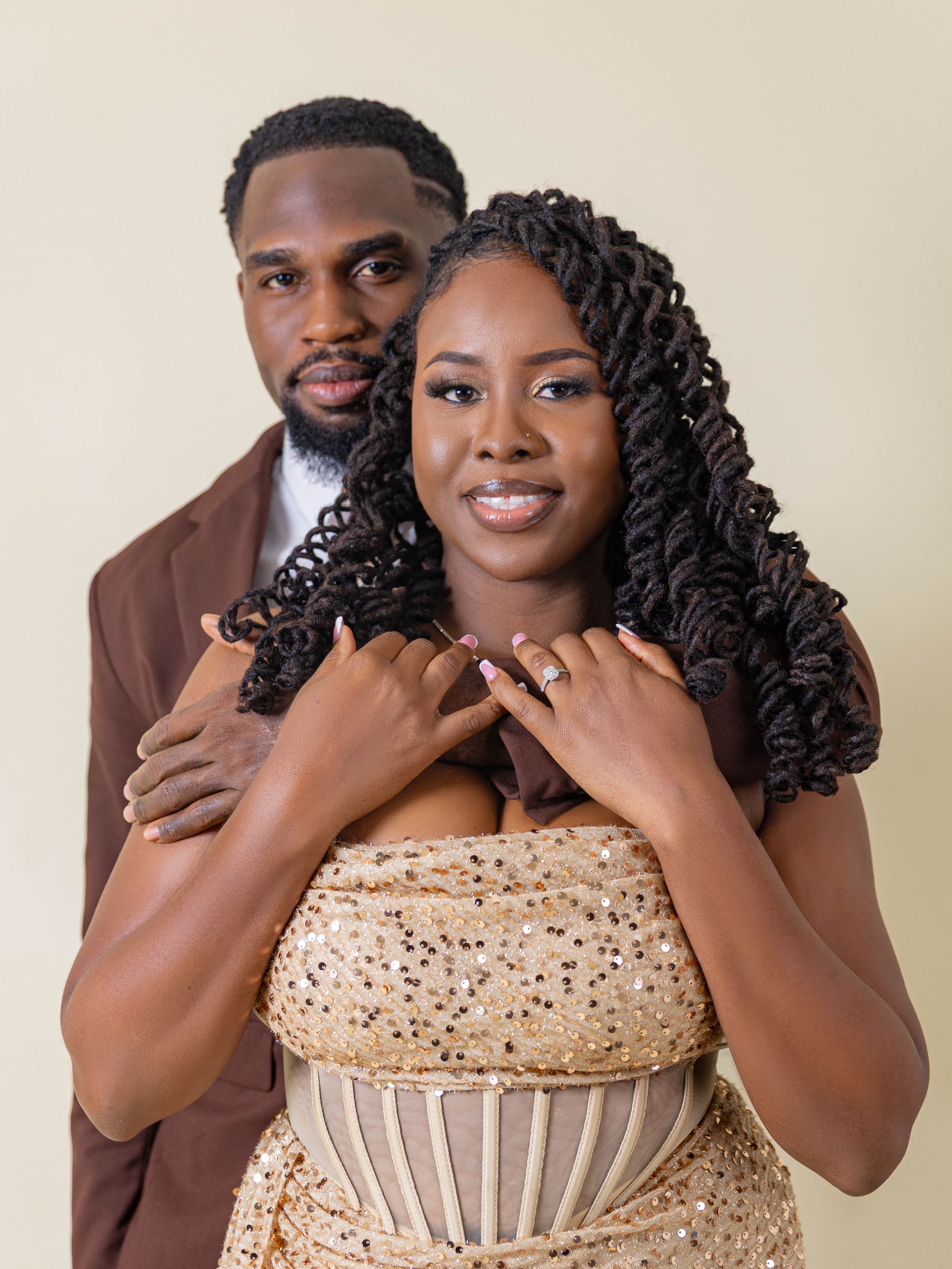 A couple in formal wear posing together, woman displaying an engagement ring while man embraces her from behind.