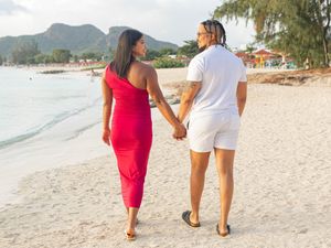 Couple holding hands walking on tropical beach, woman in red dress and man in white outfit.