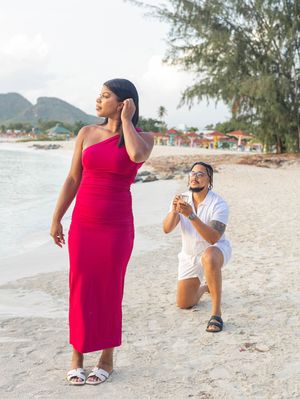 Man proposing on one knee to woman in red dress on tropical beach with ring box.