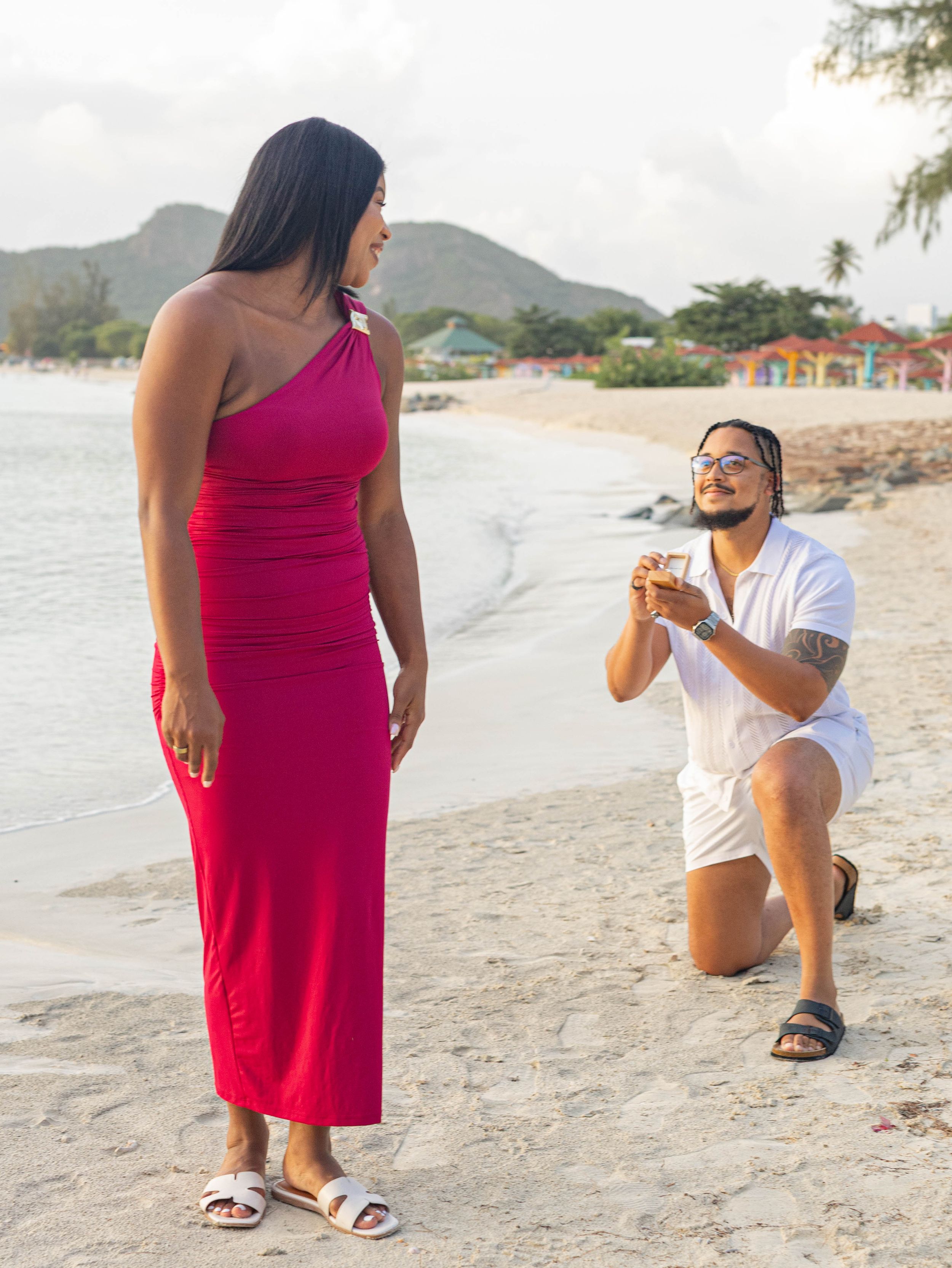 Man proposing on one knee to woman in red dress on tropical beach with mountains in background.