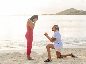 Man proposing on one knee to woman in red dress on tropical beach at sunset.