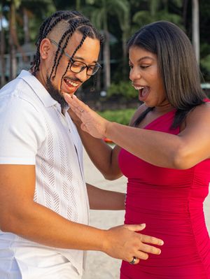 A joyful pregnant couple poses outdoors, woman in red dress touching man's face, both laughing near tropical trees.