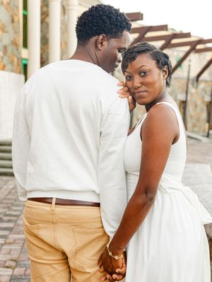 A couple dressed in white holds hands outdoors, the woman glancing back at the camera with a serene smile.