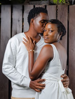 A couple in white outfits share a tender embrace, the man kissing the woman's forehead in a romantic pose.
