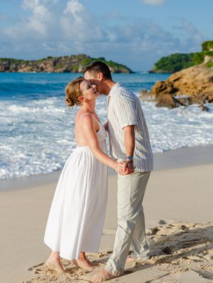 A couple holds hands and shares a romantic moment on a tropical beach with turquoise waves and rocky cliffs behind them.