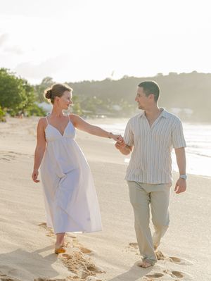 A couple walks hand-in-hand on a sandy beach at sunset, woman in white dress, man in striped shirt and khaki pants.
