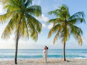Couple embracing on a tropical beach between two palm trees with turquoise ocean waves in the background.