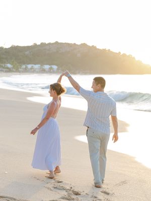 Couple dancing on a sandy beach at golden sunset, woman in white dress being twirled by man in striped shirt.
