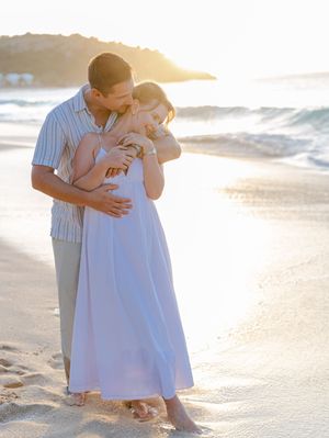 Couple embracing on a sandy beach at golden sunset, woman in white maxi dress, waves in background.