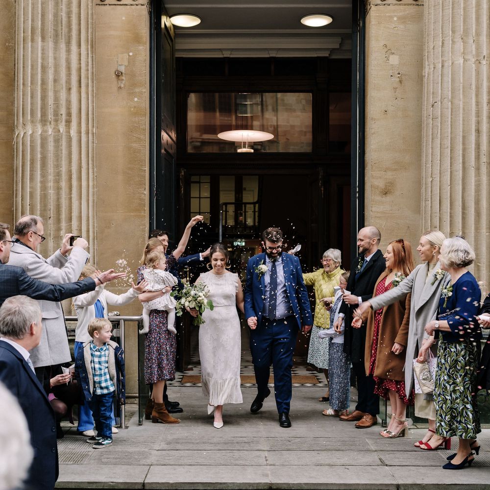 Newlyweds exit Corn Street Registry Office Wedding, Bristol as guests throw confetti in a joyful wedding celebration.