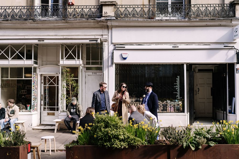 Elegantly dressed wedding party walks past charming Parisian shopfronts surrounded by spring flowers for Corn Street Registry Office Wedding