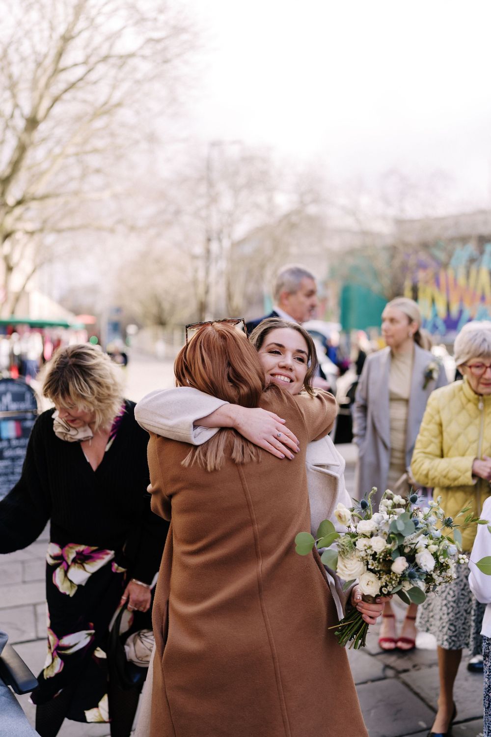 Bride holding bouquet hugs a guest in brown coat outdoors at a sunny wedding celebration at Corn Street Registry Office Wedding, Bristol