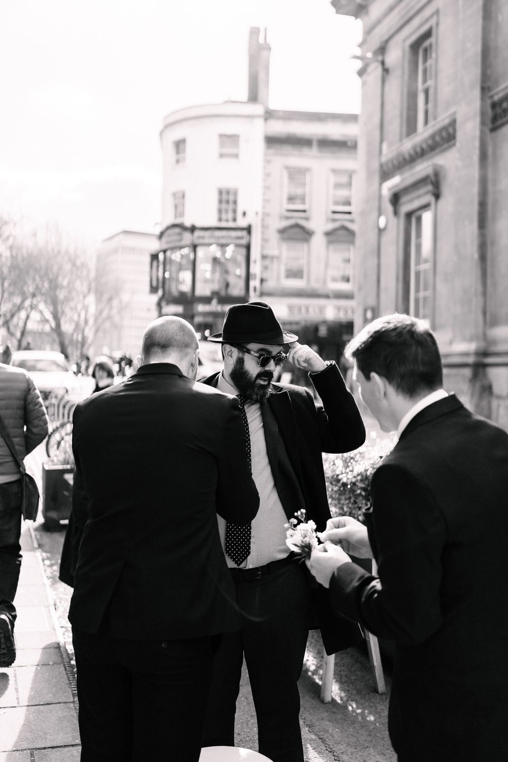 Black and white photo of men in suits on a city street, one wearing a hat and sunglasses at Corn Street Registry Office Wedding, Bristol