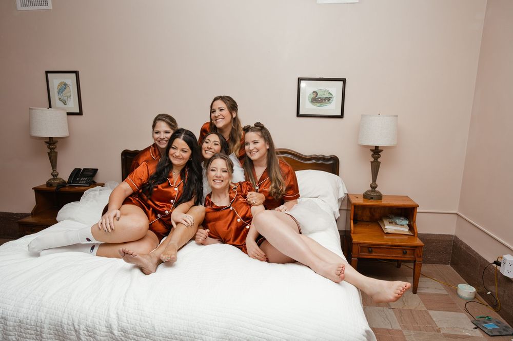Six bridesmaids in matching rust satin robes laughing together on a white bed during wedding morning preparations.