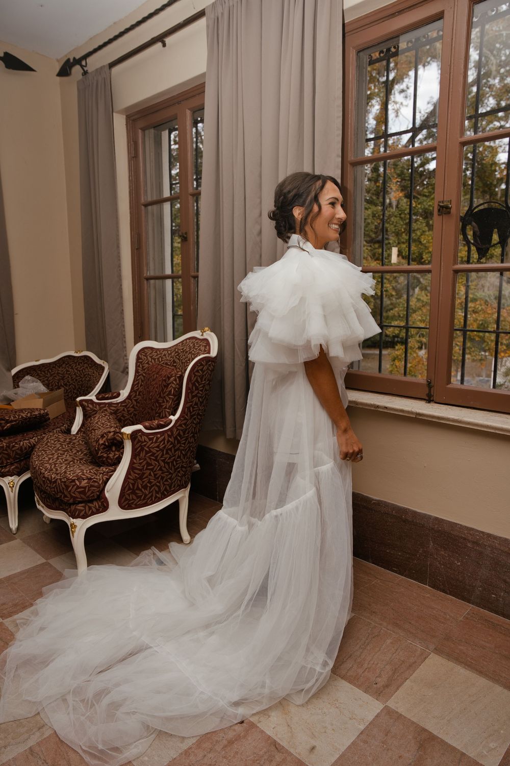 Bride in elegant white tulle wedding gown with ruffled collar and flowing train, smiling in a decorated room.