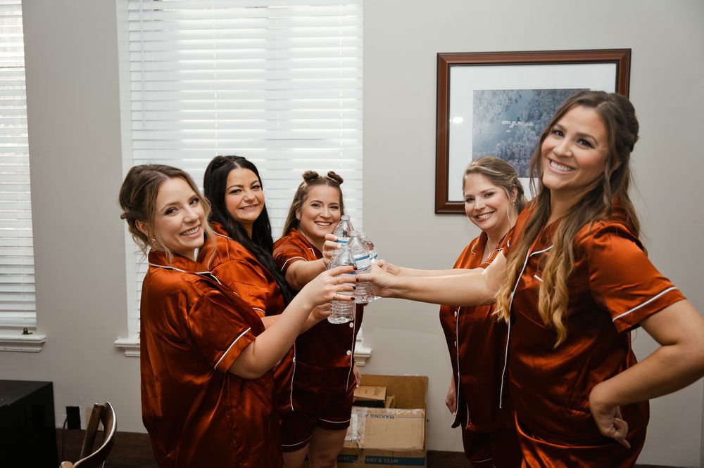 Five bridesmaids in rust velvet robes toasting with a water bottle while getting ready for a wedding.