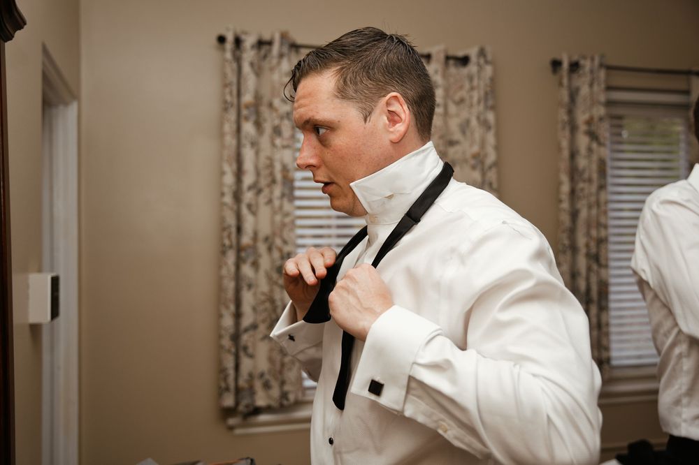 Groom in white tuxedo jacket adjusting black bow tie while getting ready for wedding ceremony.